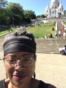 On a trip of the decade, Black woman with glasses and headband in front of the steps leading to the Sacre-Coeur, the white building in the background
