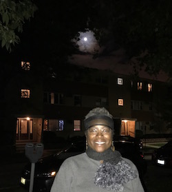 Hunters moon above a Black woman in gray shirt and gray scarf standing in front of a building